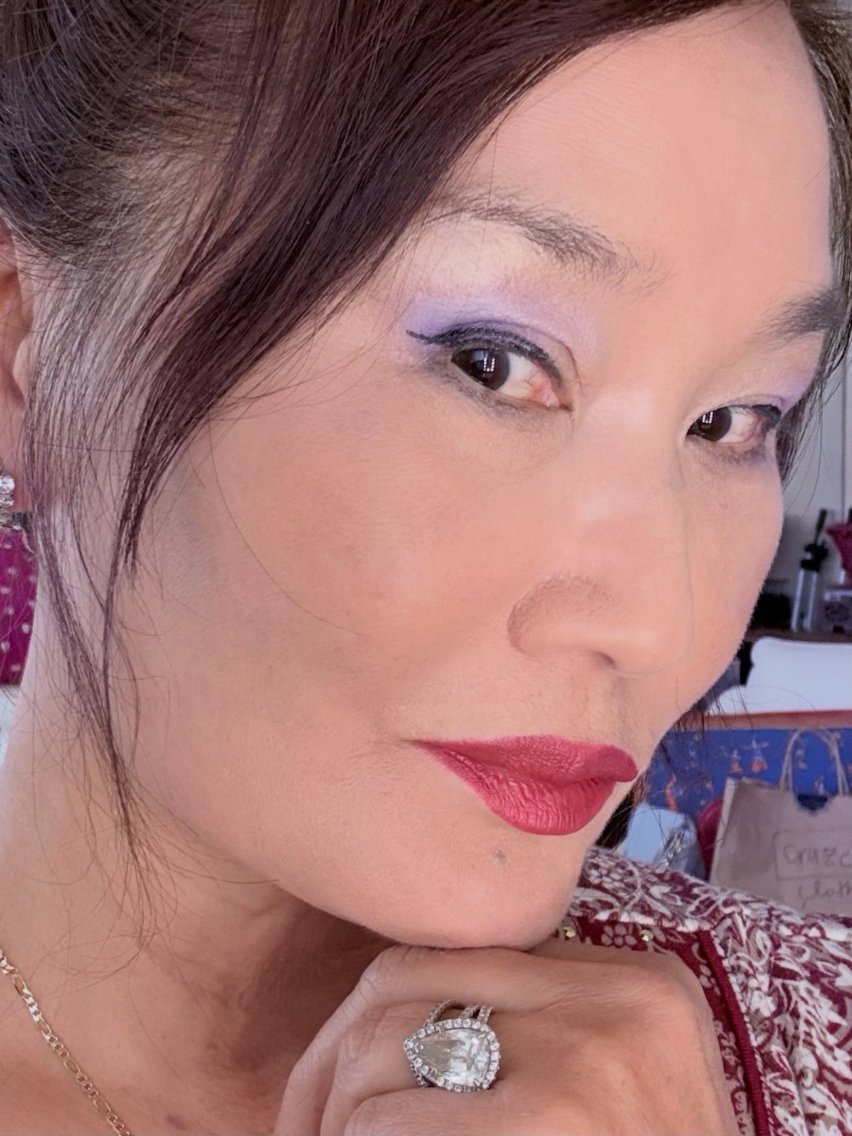 Woman with bold red lips, purple eyeshadow, and a large diamond ring.