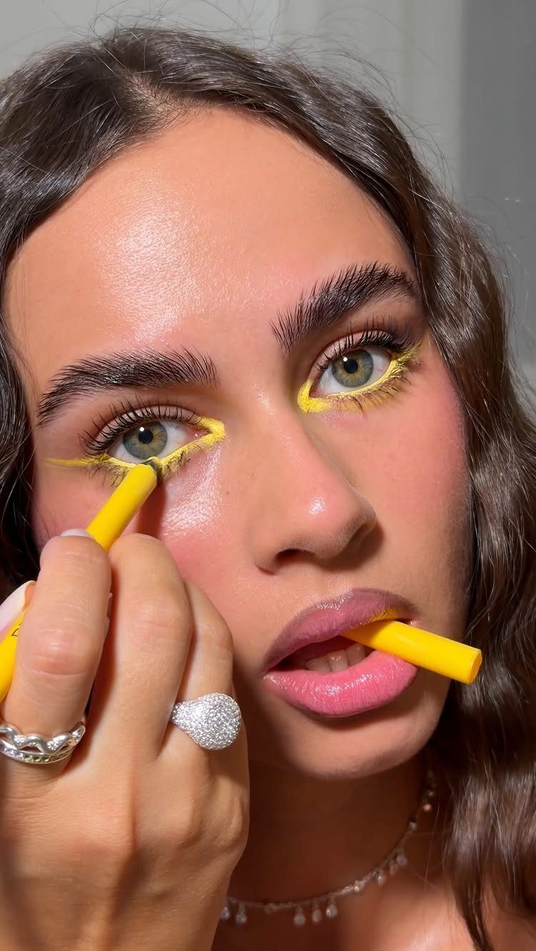 Woman applying bright yellow eyeliner to her lower eyelids with a yellow pencil.