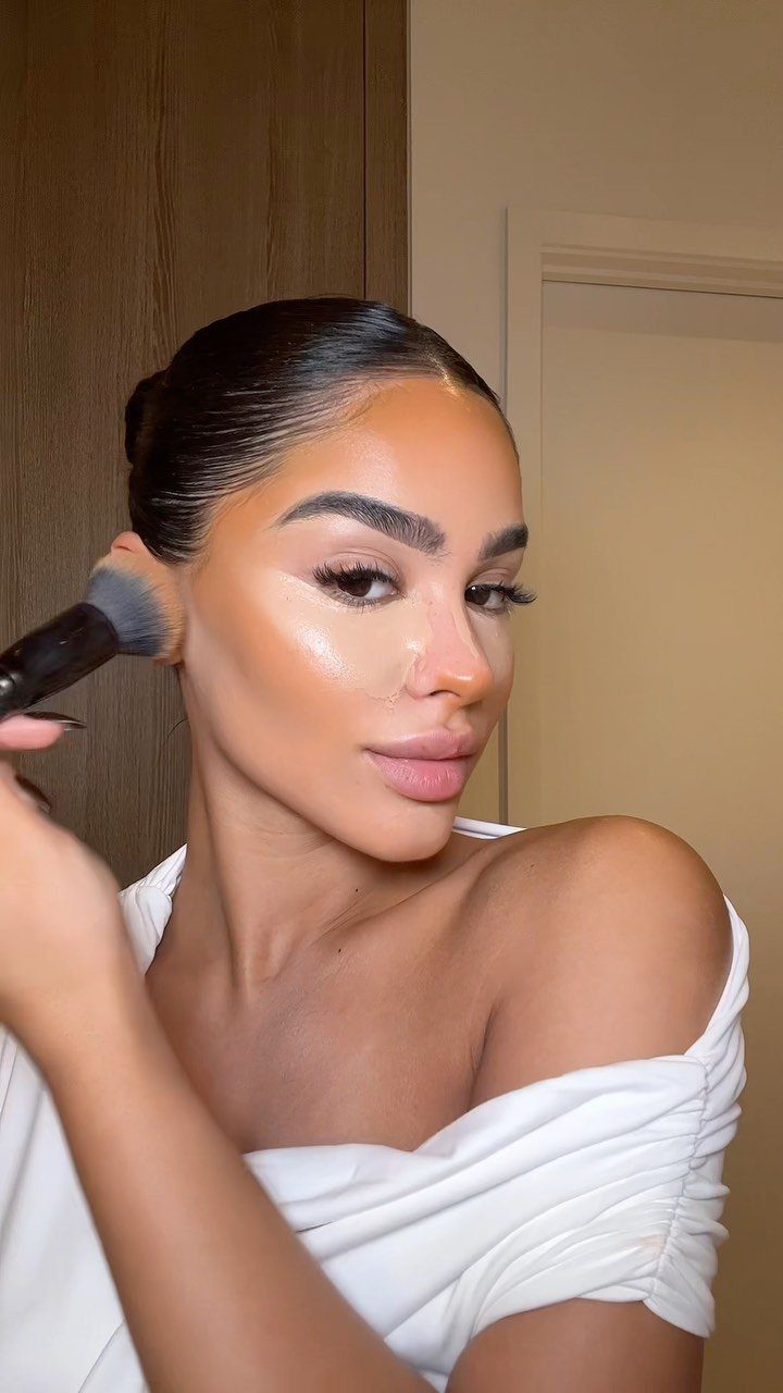 Woman applying makeup to her cheek with a brush, featuring glowing highlighter.