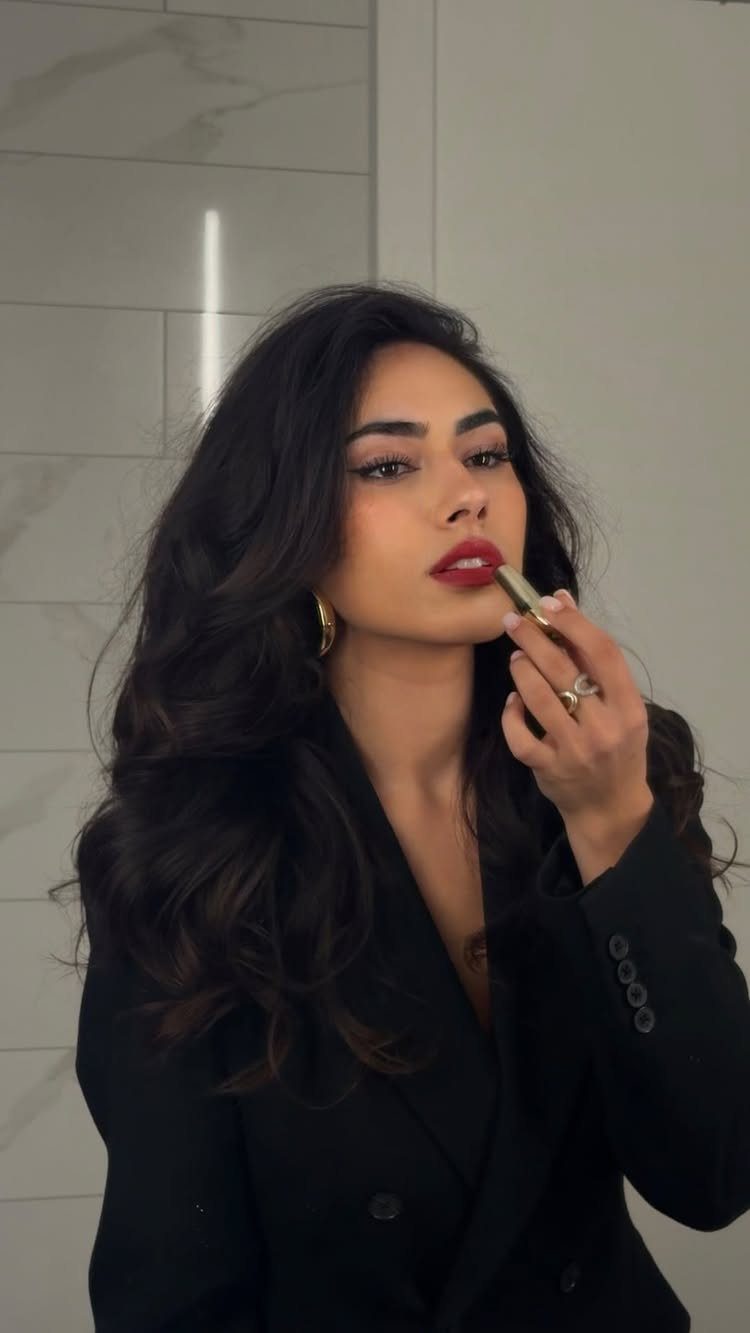 Woman with elegant makeup applying red lipstick while wearing a black blazer.