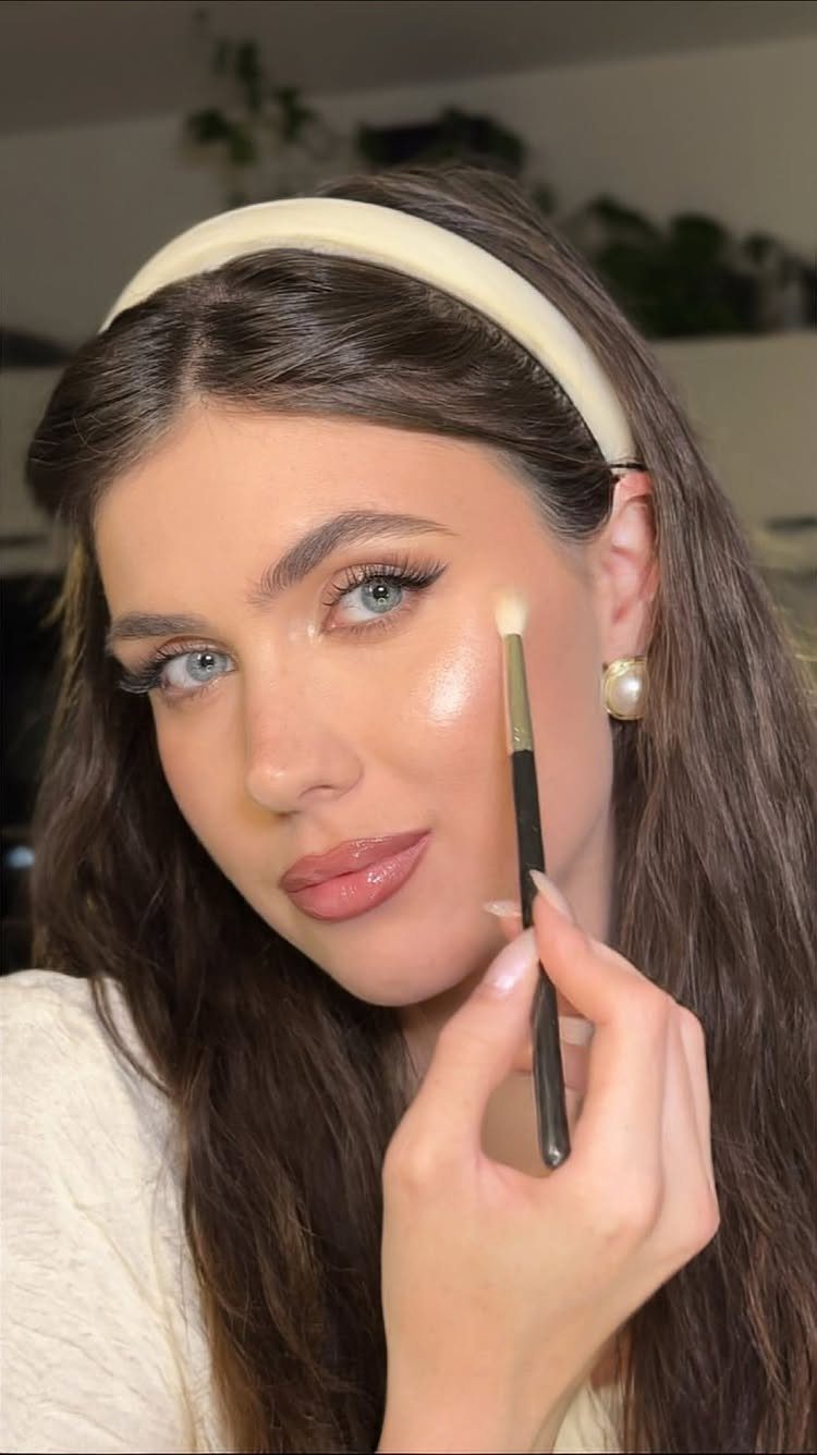 Woman applying creamy highlighter to her cheek with a makeup brush for glowing skin.