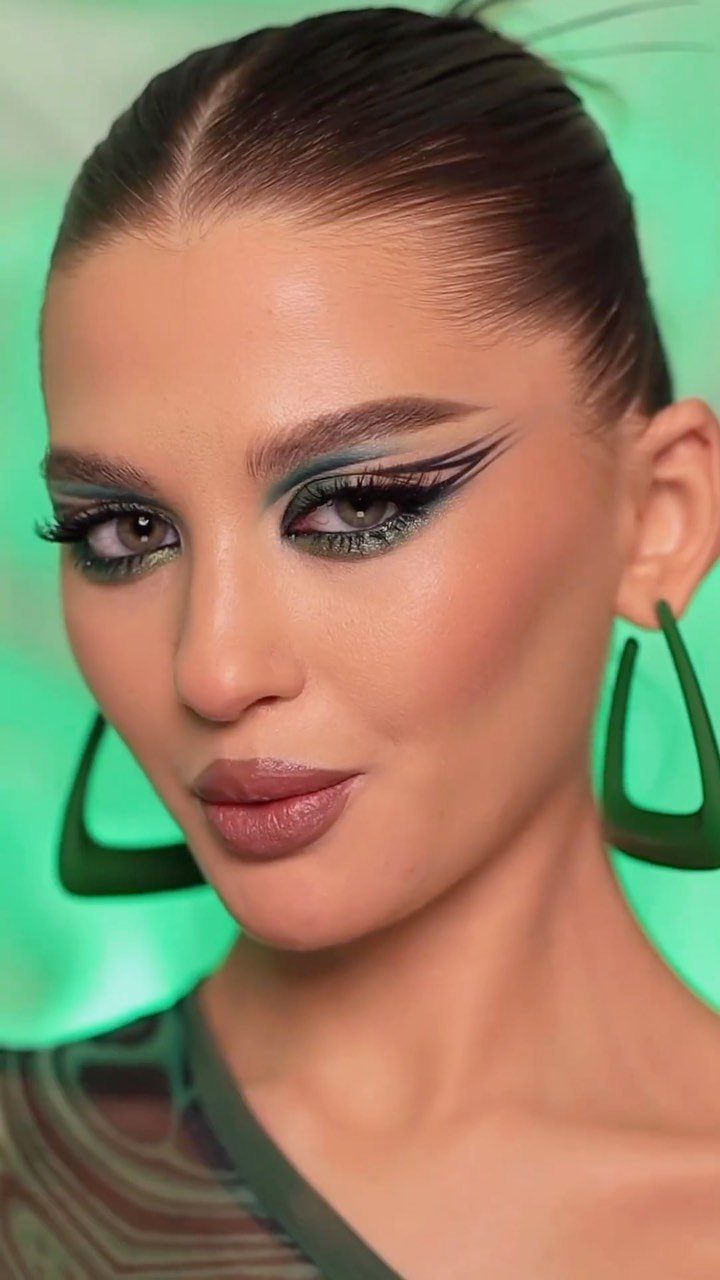 Woman with bold green winged eyeliner and matching geometric earrings against green background