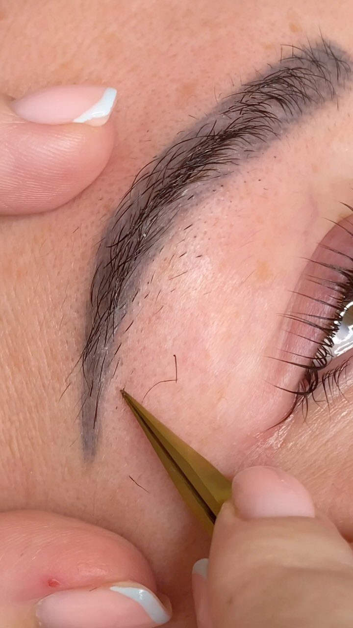 Gold tweezers plucking a stray hair from below a shaped dark eyebrow.