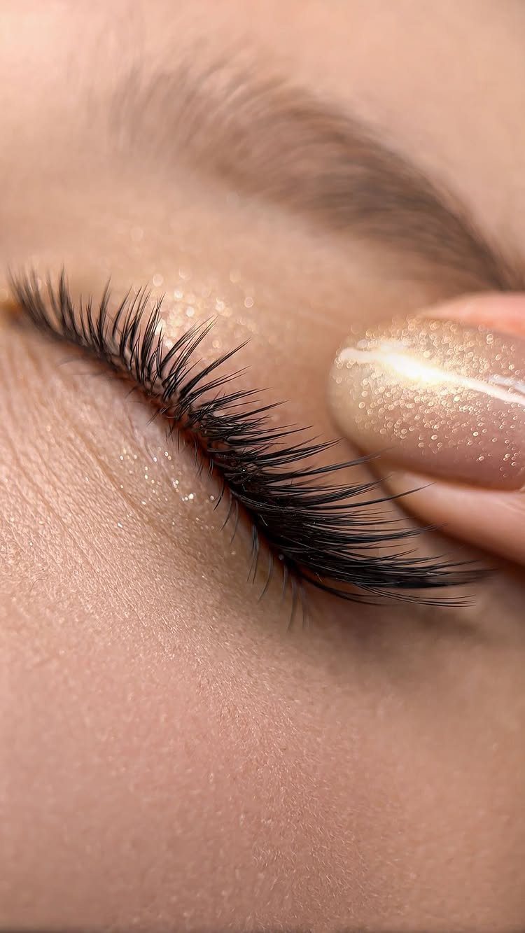 Long dark eyelashes being lifted by a finger with gold glitter nail polish.