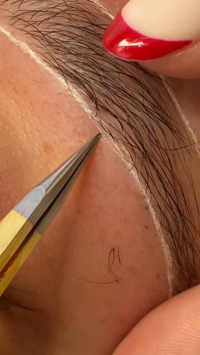 Tweezers plucking a stray hair from an eyebrow outlined in white makeup.