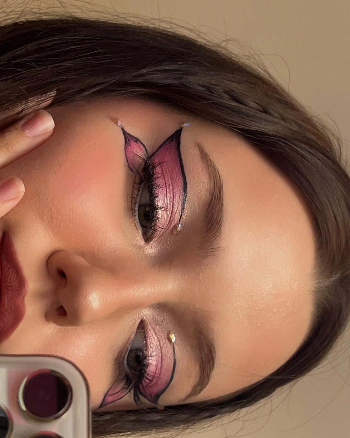 Person with pink eyeshadow and bold black butterfly-shaped graphic eyeliner and pearl accents.