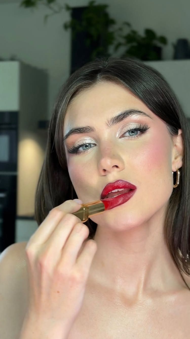 Woman applying bold red lipstick with shimmering silver eyeshadow and winged eyeliner.
