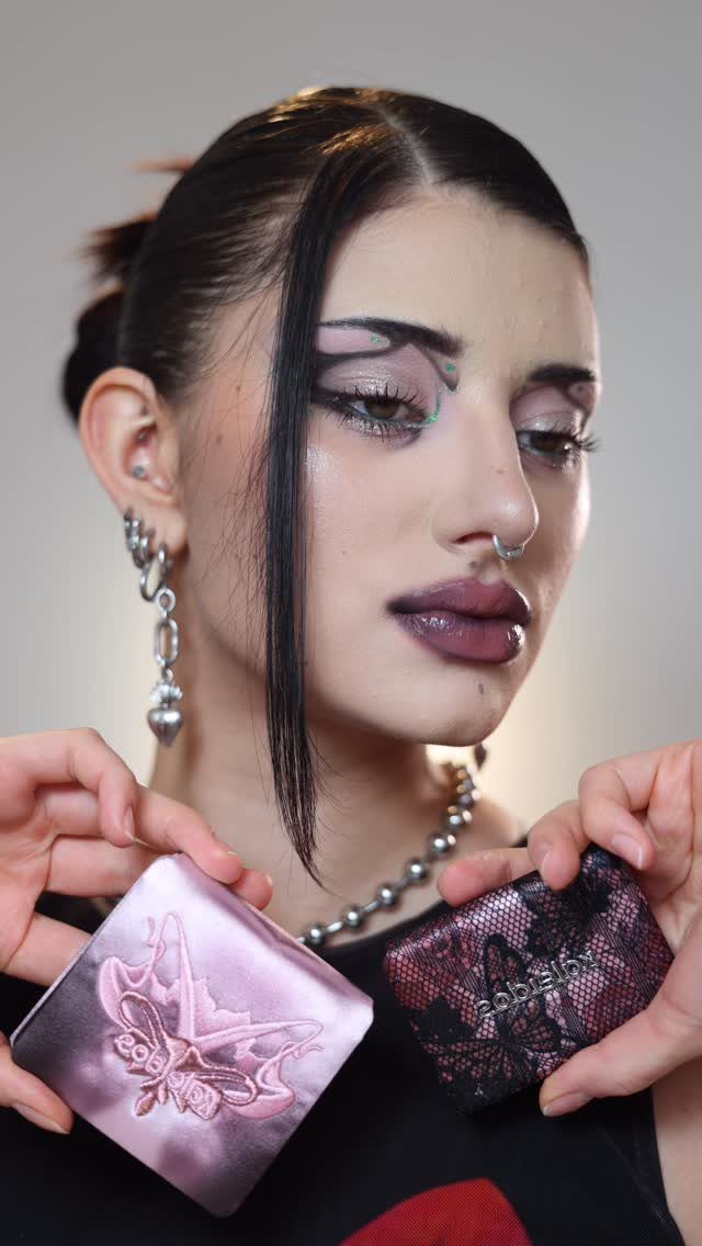 Woman with graphic black eyeliner and dark purple lips holding two makeup compacts.