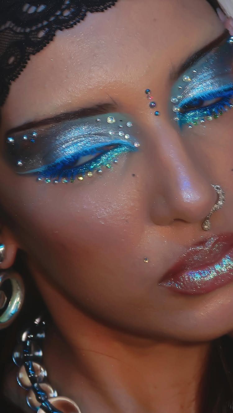 Person with shimmering blue eyeshadow, rhinestones around eyes, and iridescent holographic lipstick.