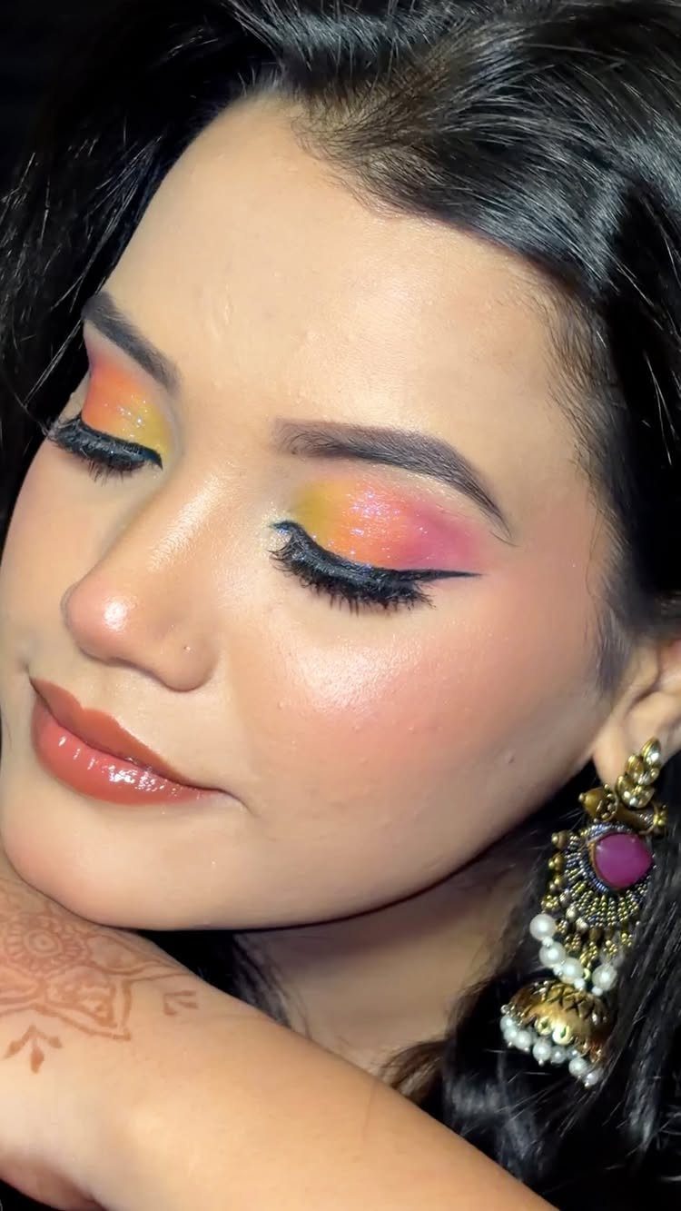 Woman with colorful orange and pink eyeshadow, winged eyeliner, and traditional gold earrings.