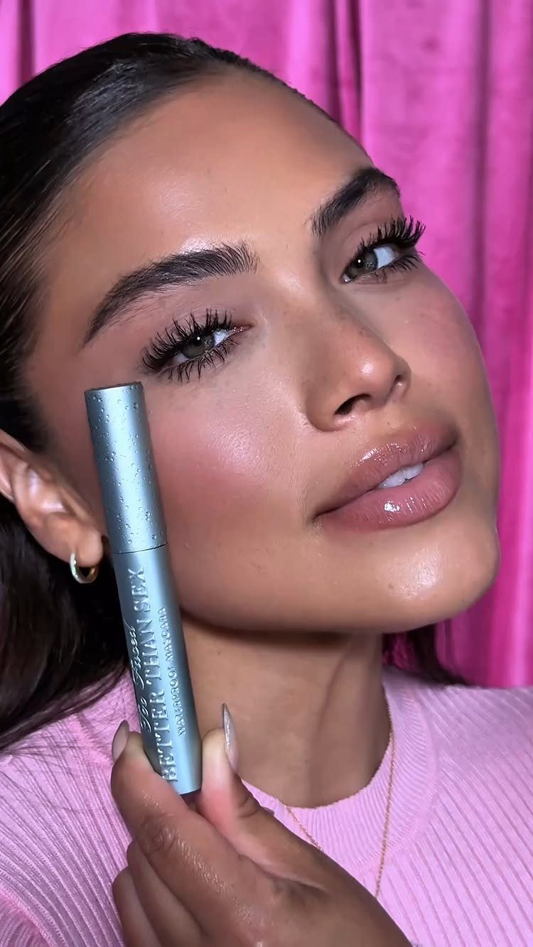 Woman with voluminous dark eyelashes holding a silver mascara tube against her cheek.