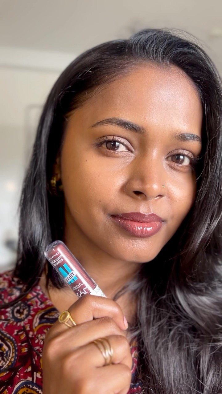 Woman with brown skin wearing berry lip color and holding a lip gloss tube.