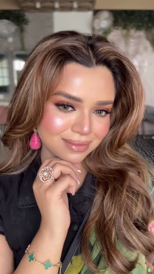 Woman with brown skin wearing shimmering makeup, pink blush, and wavy brown hair.