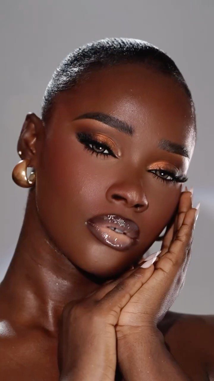 Woman with dark skin wearing gold eyeshadow, glossy lips, and radiant highlighter.