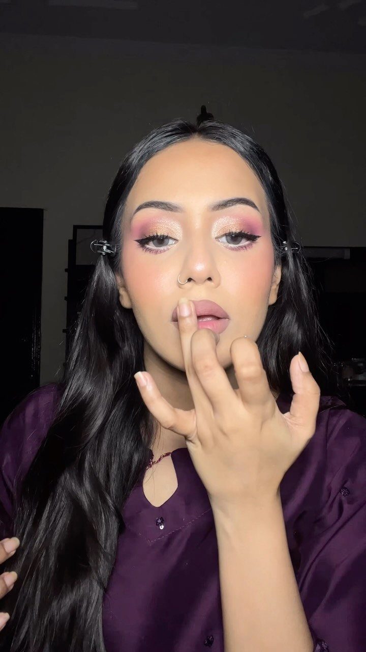 Woman with dark skin wearing pink and gold eyeshadow and purple clothing.