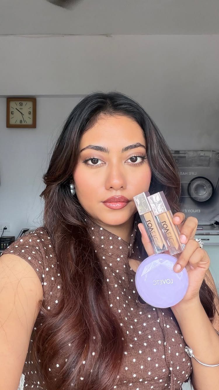 Woman with soft glam makeup holding foundation bottles and a purple compact.