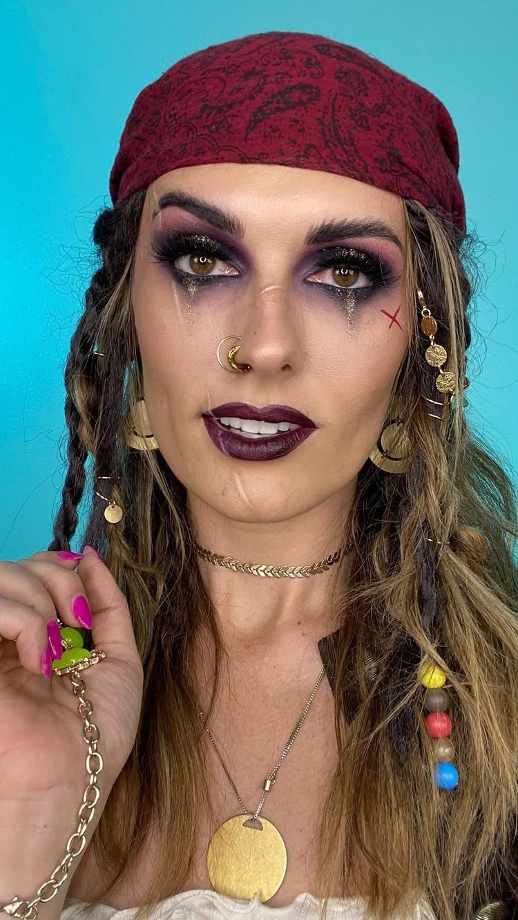 Woman with pirate makeup, red bandana, and gold jewelry posing against blue background