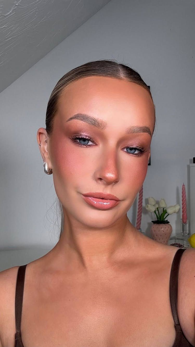 Woman with rose gold shimmering eyeshadow, pink blush, and glossy lips posing indoors.