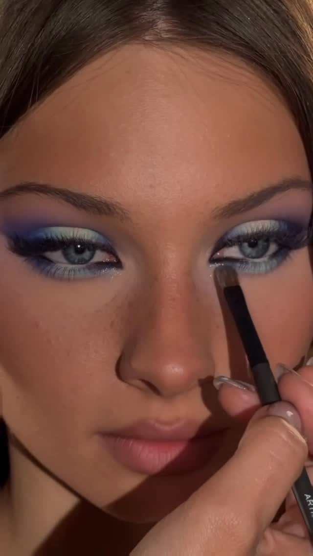 Woman with blue eyes wearing purple eyeshadow while a brush applies makeup.