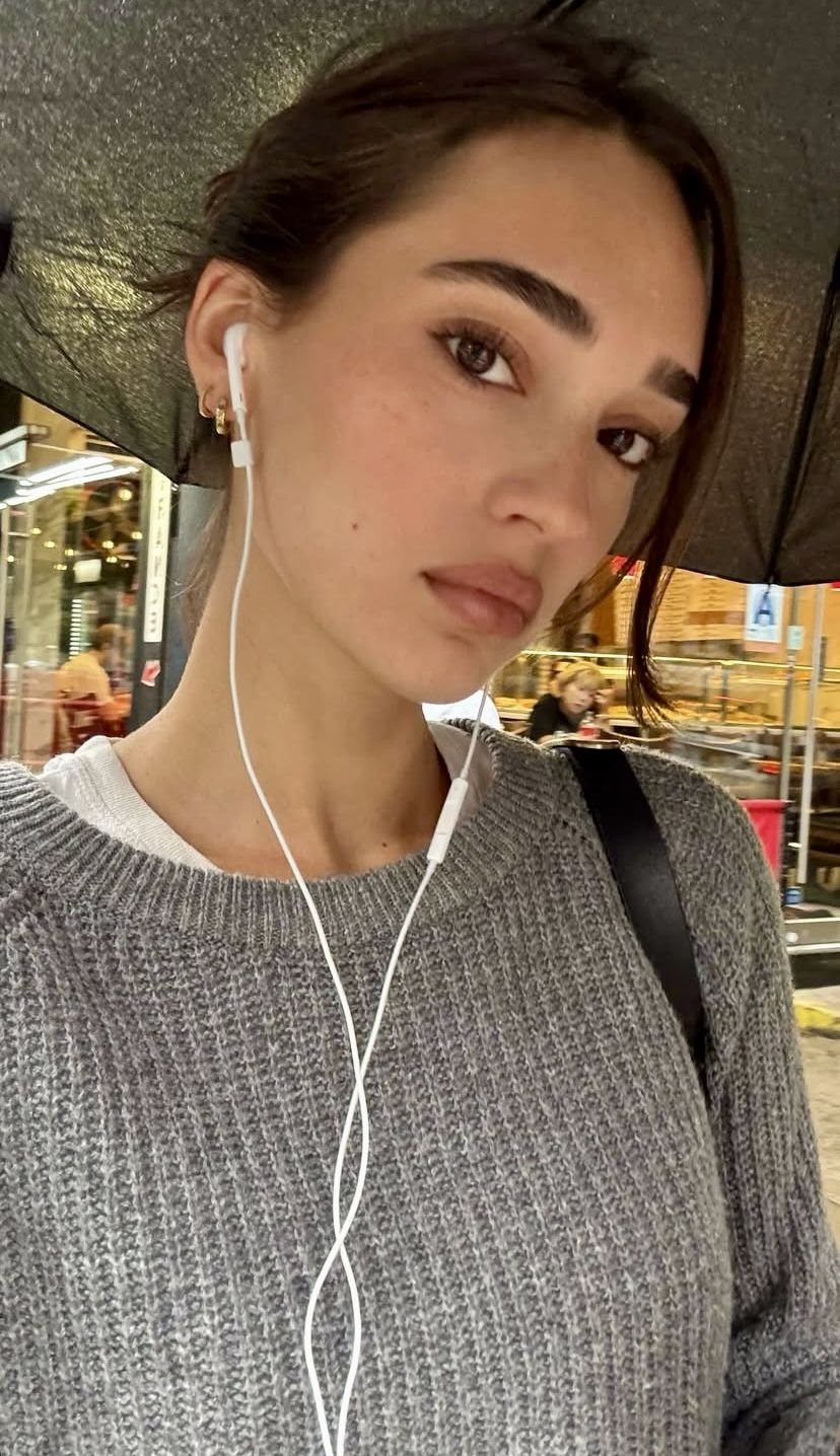 Woman with minimal makeup wearing a grey sweater under a black umbrella.