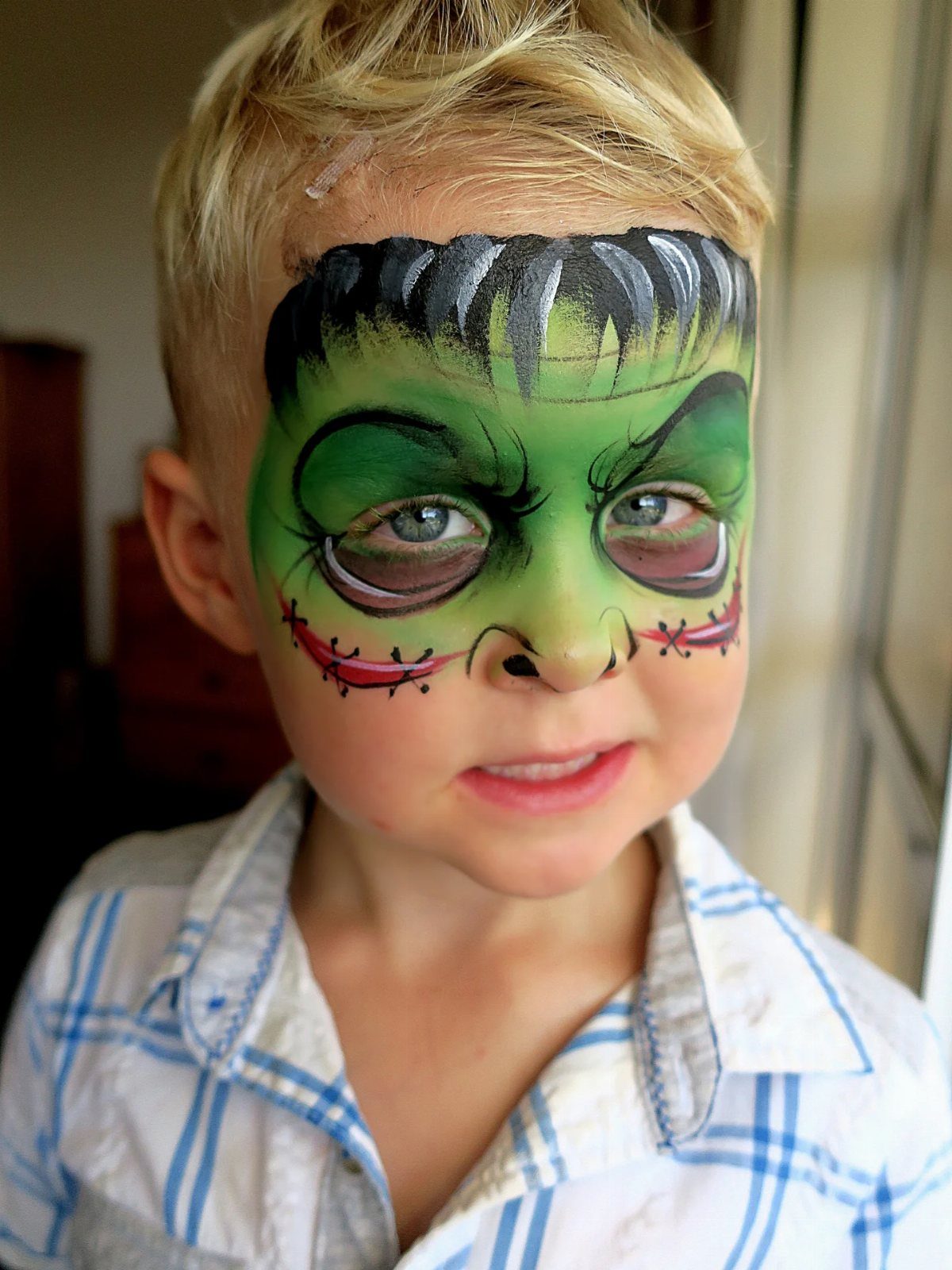 Young boy with green Frankenstein face paint and red stitched cheek details.