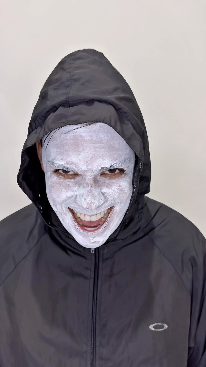 Person with white ghost face paint smiling while wearing a black hooded jacket.