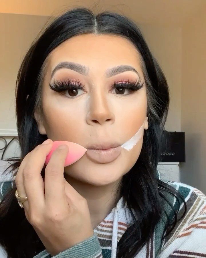 Woman applying foundation with a pink sponge, featuring soft glam makeup and lashes.