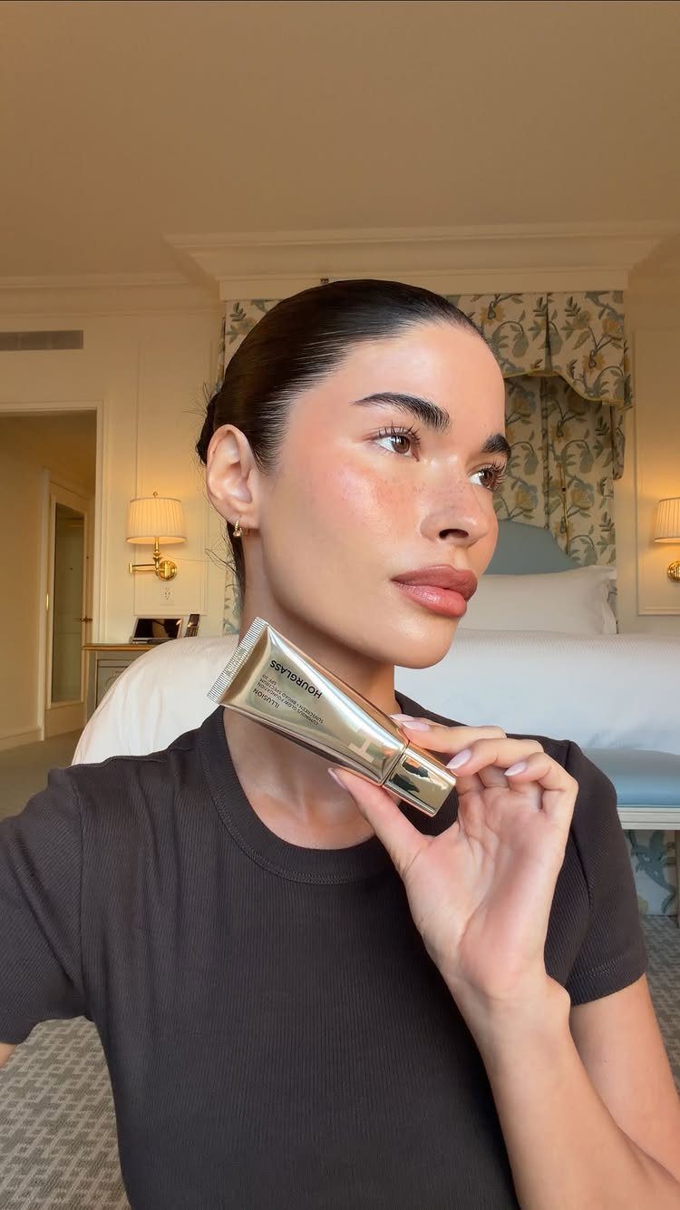 Soft Sultry Makeup Ideas: 10 Looks for Day and Night 1 Woman with glowing skin holding a gold tube of liquid foundation in a bedroom