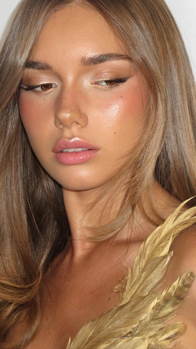 Woman with winged eyeliner and glossy pink lips wearing gold feathered clothing.