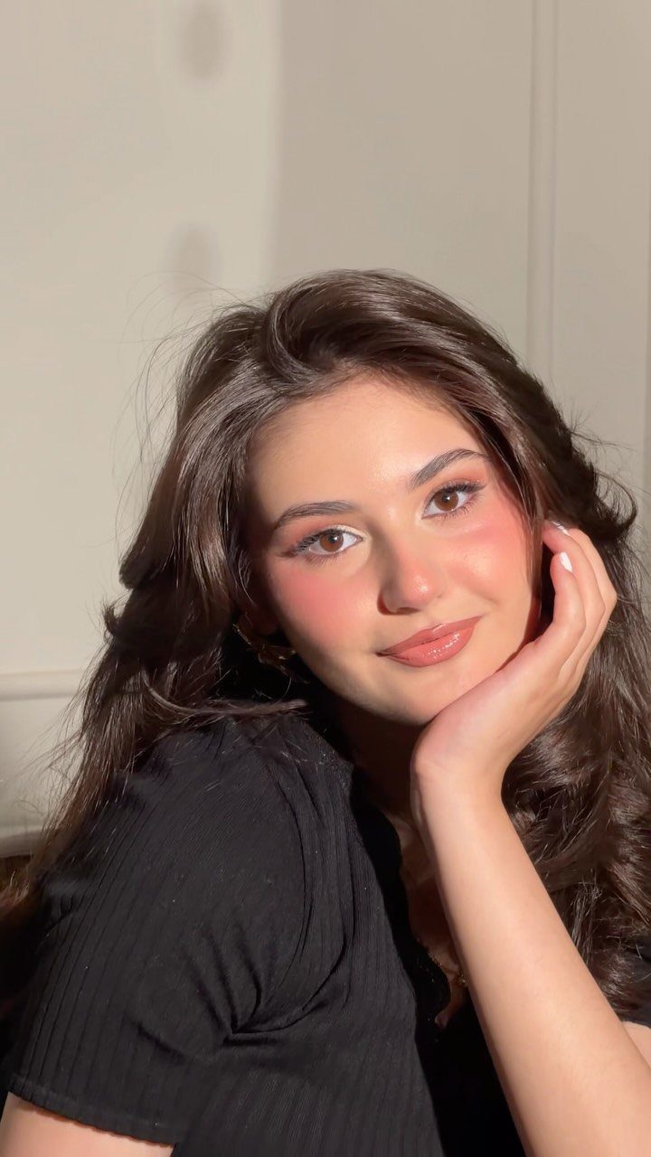 Young woman with soft glam makeup and wavy brown hair posing against white wall