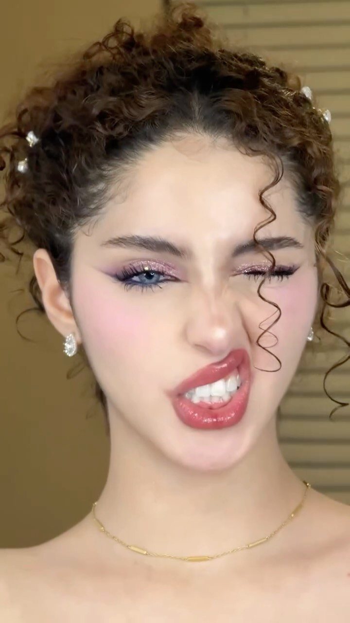 Woman with curly hair wearing glossy red lips and shimmery purple eyeshadow.